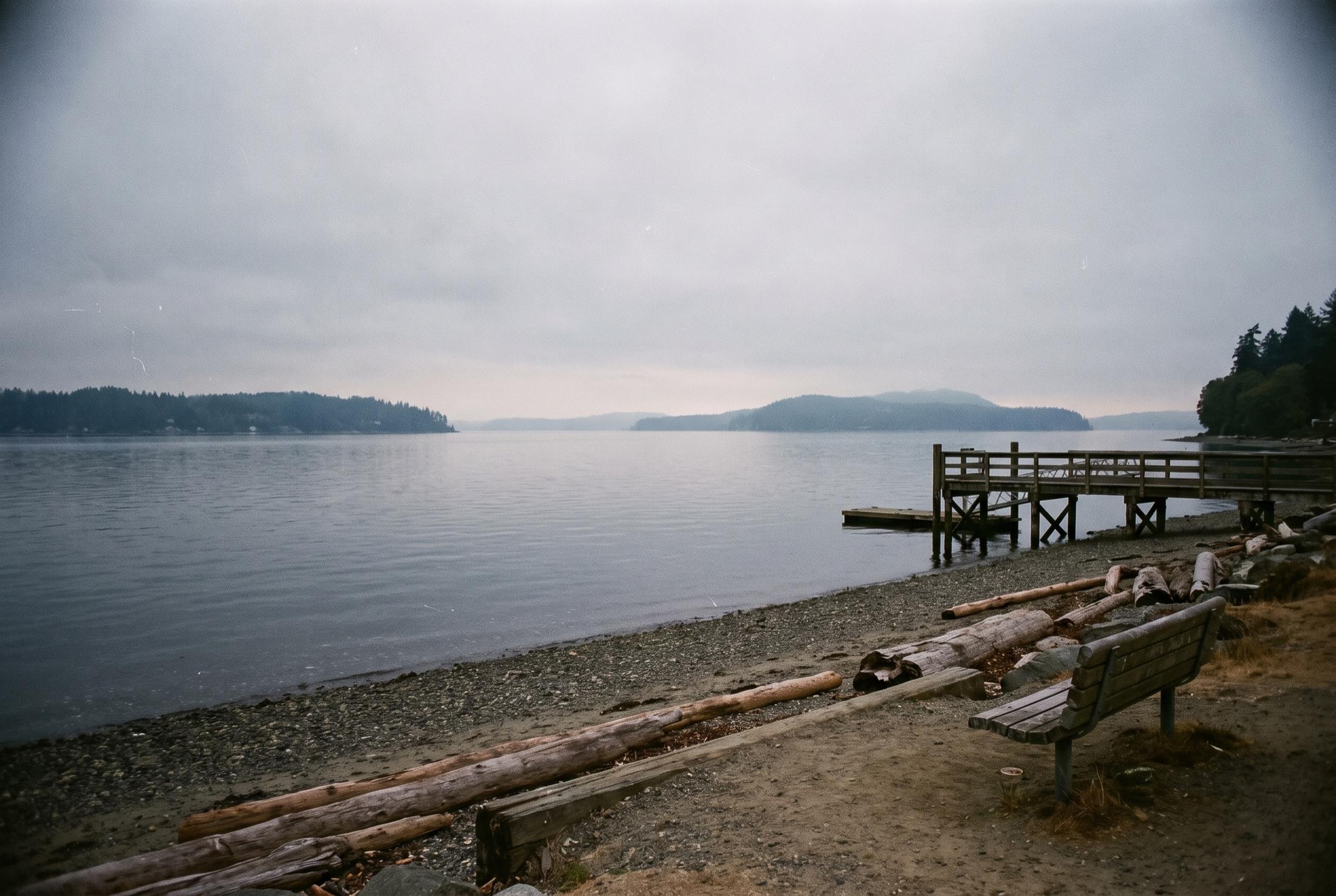 Gravel beach on Puget Sound under an overcast sky, with driftwood logs, a wooden bench, a small pier, and forested islands across calm gray water
